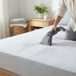 Une personne nettoie un matelas blanc avec une shampouineuse, laissant des traces d'eau savonneuse. Chambre lumineuse et propre.